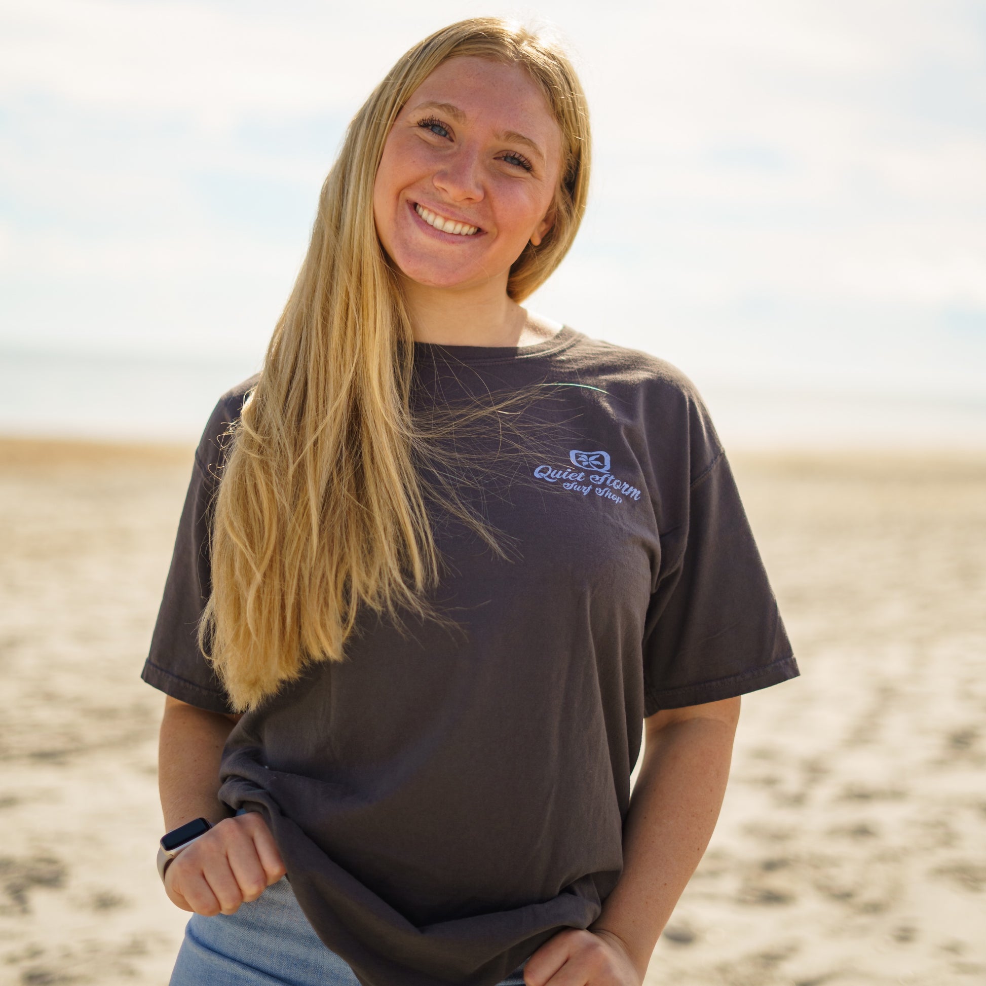 Women wearing Front of grey T-shirt with a small graphic with a Q and palm tree inside it with Quiet Storm Surf Shop underneath it