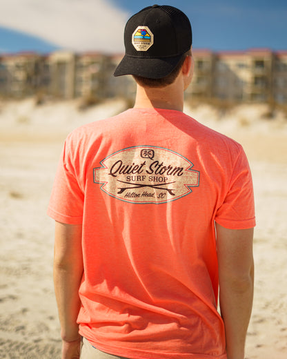Man at beach wearing a hat and Graphic T shirt that has a design of of Quiet Storm Surf shop with two surf boards crossing underneath with Hilton Head, SC below