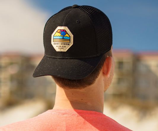 Showing a Man wearing a hat that shows the front of the hat with a patch on it that has an image of a sunset with a tree and moon and below says Quiet Storm Hilton Head Island