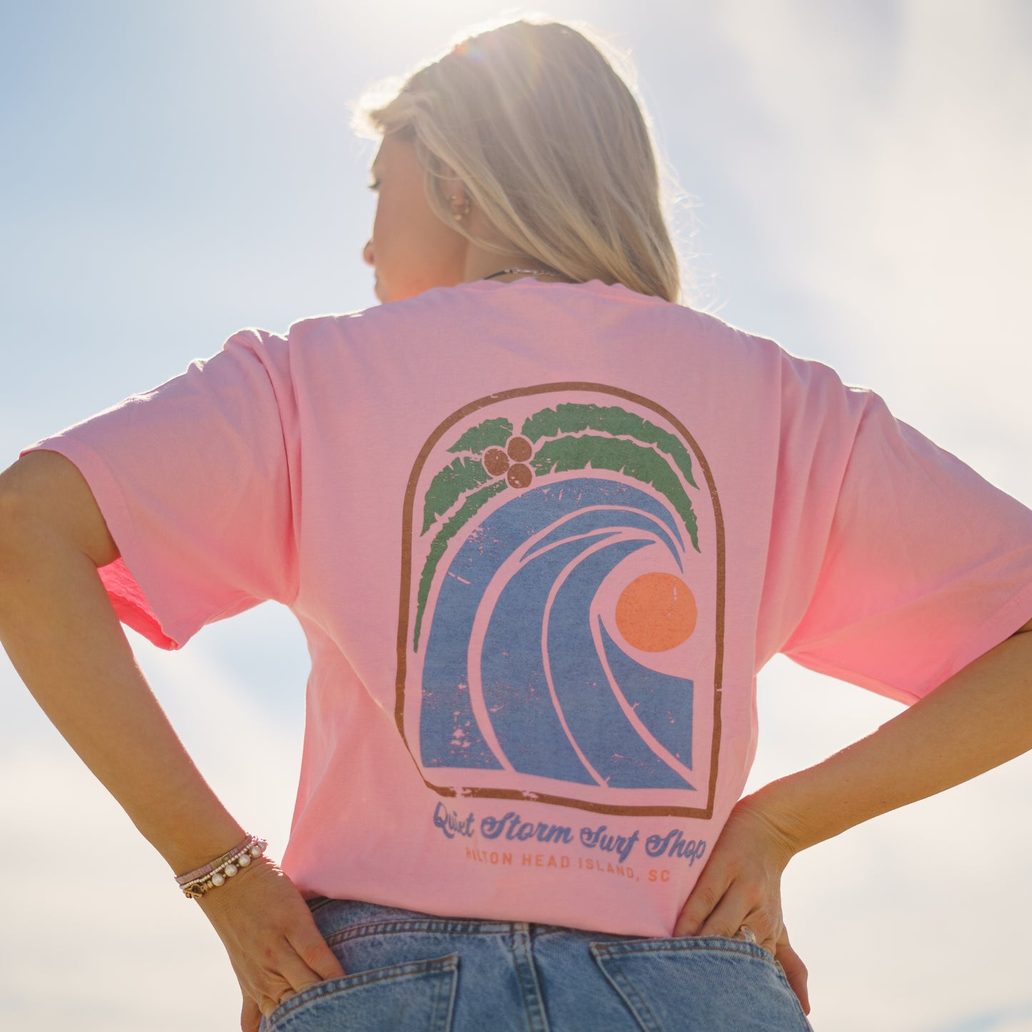 Women wearing pink T-shirt with design that has a palm tree with a wave and a moon with Quiet Storm Surf Shop Hilton Head Island, SC