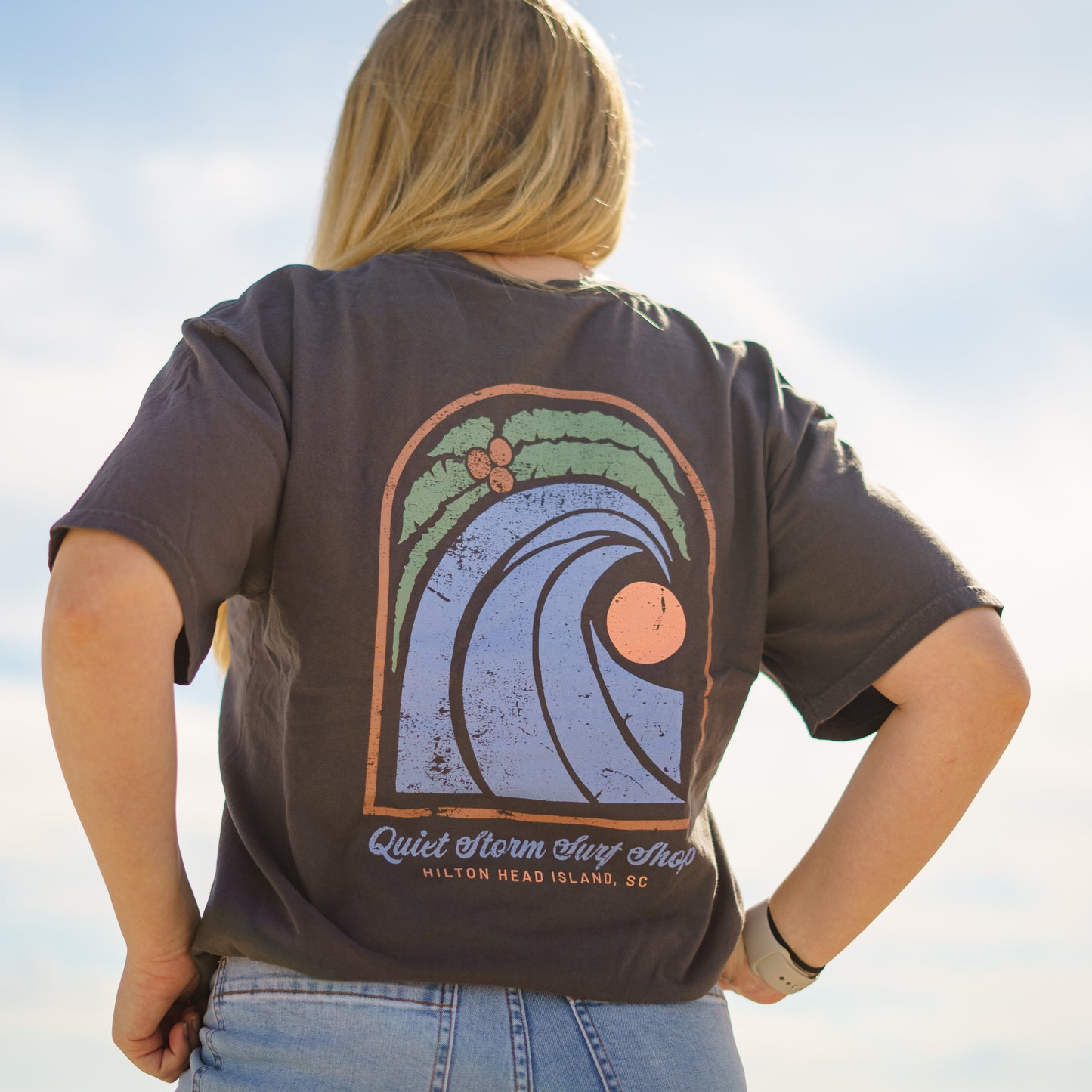 Women wearing grey T-shirt with design that has a palm tree with a wave and a moon with Quiet Storm Surf Shop Hilton Head Island, SC