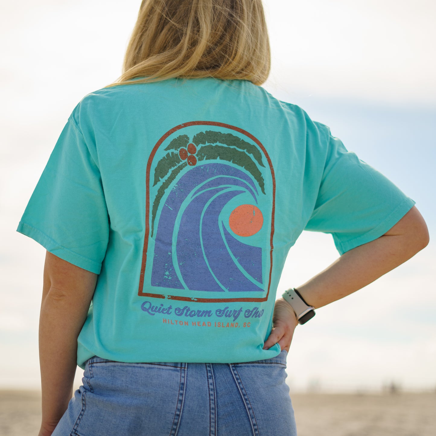Women wearing teal T-shirt with design that has a palm tree with a wave and a moon with Quiet Storm Surf Shop Hilton Head Island, SC
