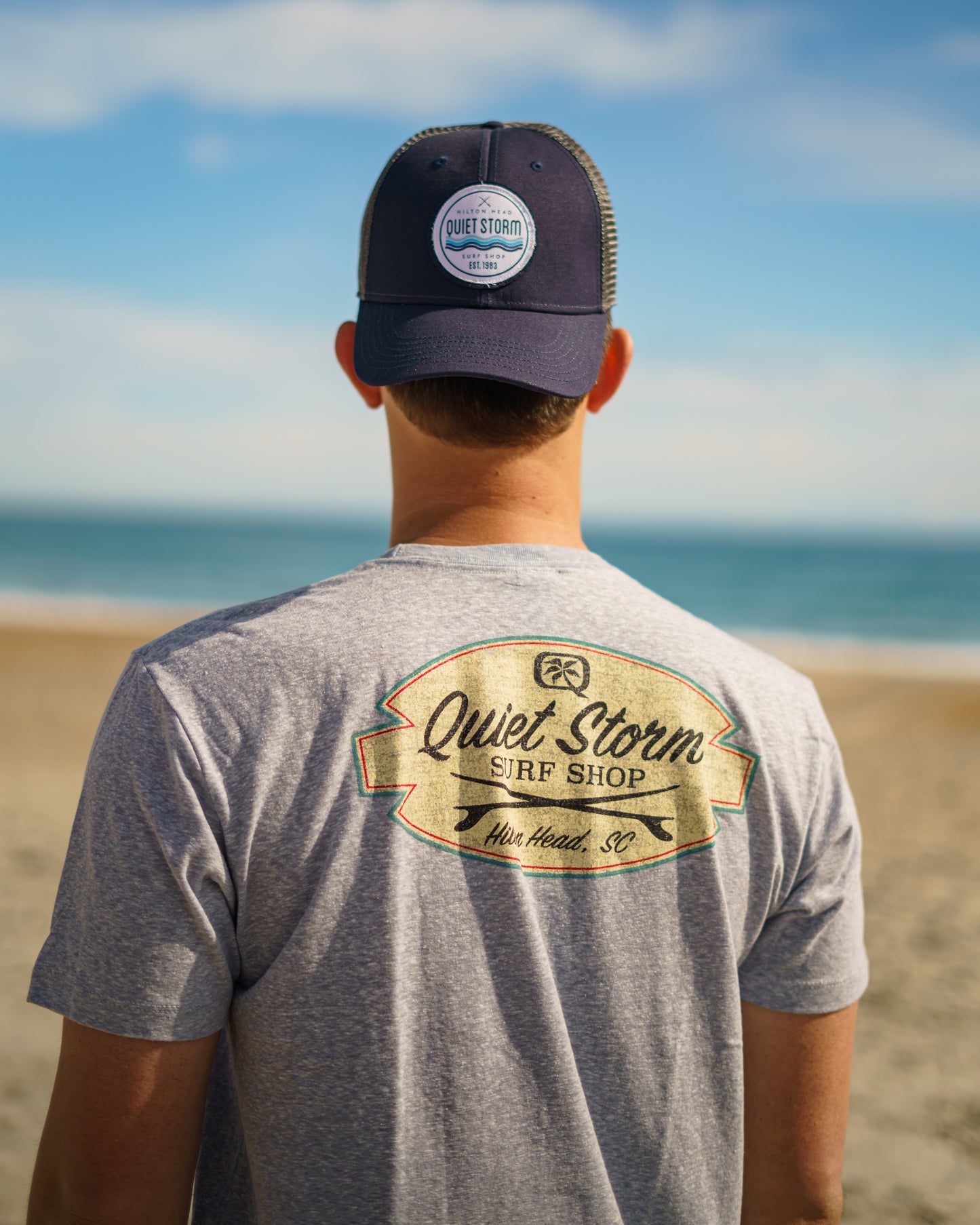 Man at beach wearing a hat and Graphic T shirt that has a design of of Quiet Storm Surf shop with two surf boards crossing underneath with Hilton Head, SC below