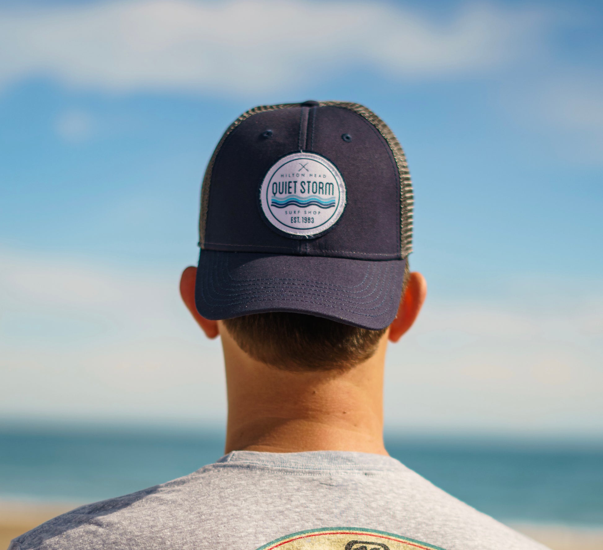 A man wearing a navy and grey hat with a mesh back and a patch featuring two crossed surfboards and the text 'Quiet Storm Hilton Head Surf Shop'."