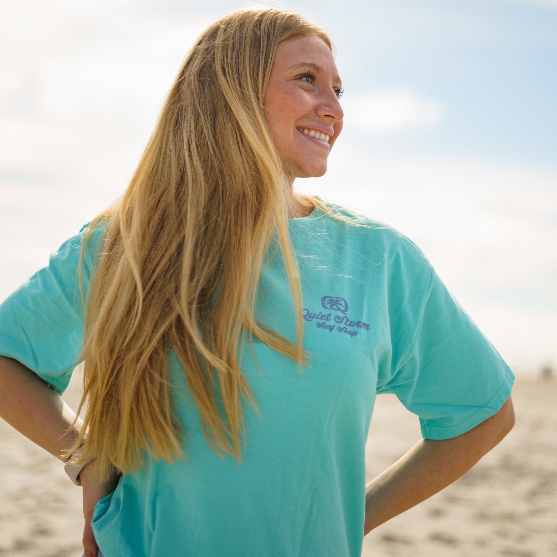 Women wearing Front of teal T-shirt with a small graphic with a Q and palm tree inside it with Quiet Storm Surf Shop underneath it  