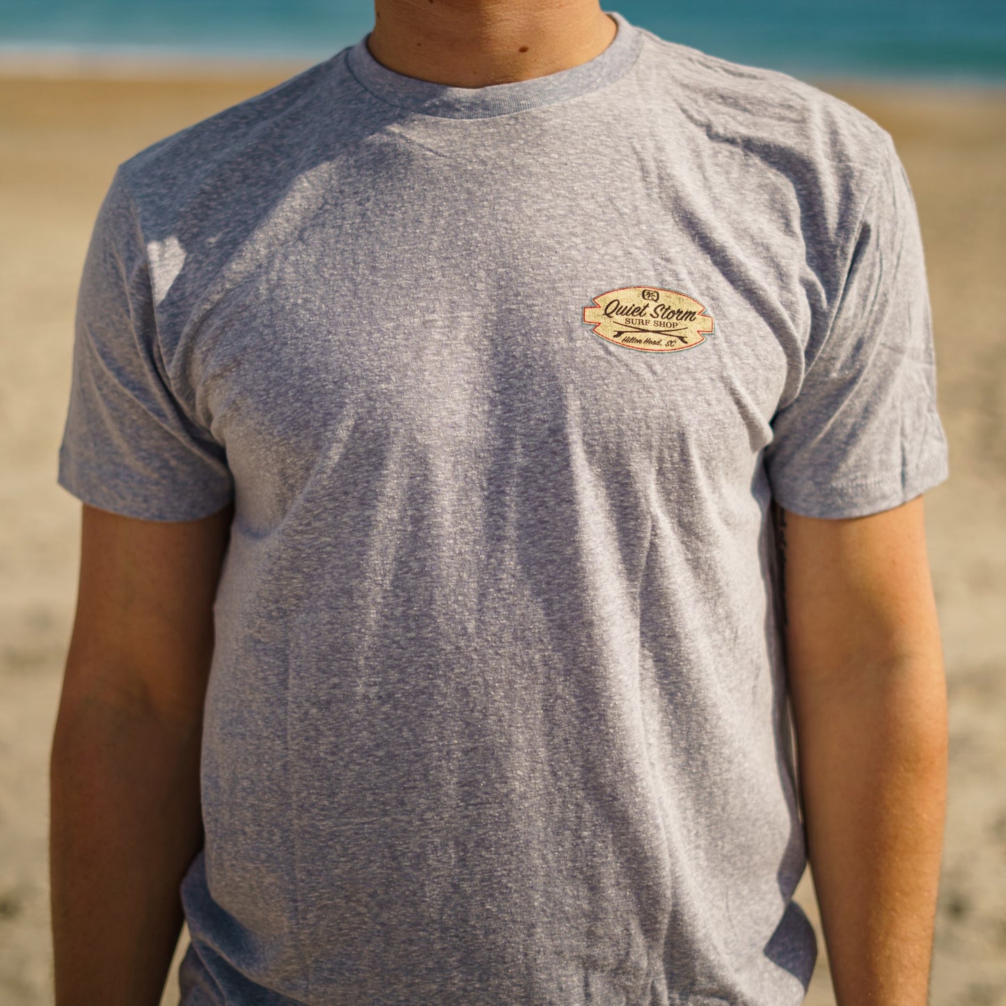 Man at beach wearing a graphic tee showing front of shirt with small design on top corner of shirt that says Quiet Storm surf shop and below has two surf boards crossing and below says Hilton Head,SC